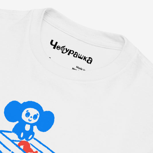 White unisex youth T-shirt with original Soviet cartoon character Cheburashka sitting on a box of oranges and チェブラーシカ logo in red. Front closeup.