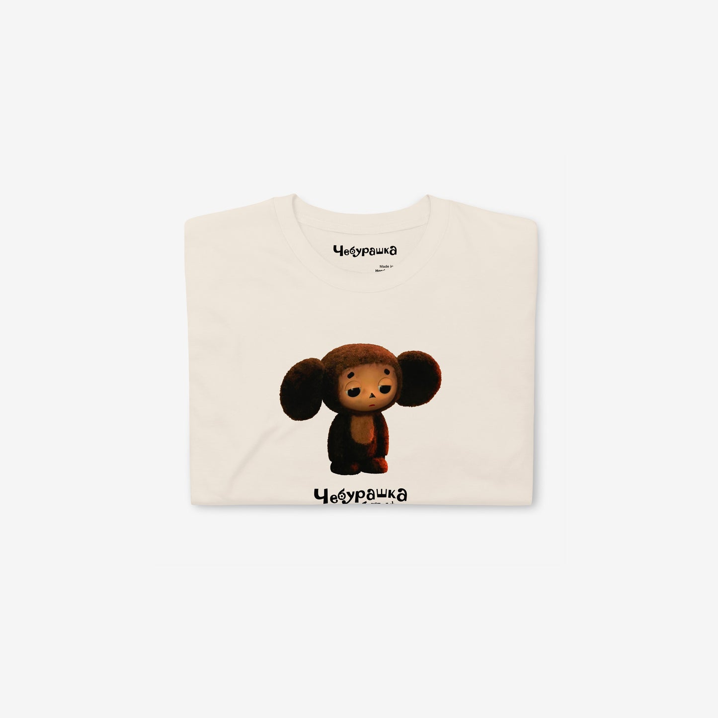Beige unisex T-shirt featuring large print of Sad Cheburashka from Soviet cartoon. With logo Чебурашка. Folded.