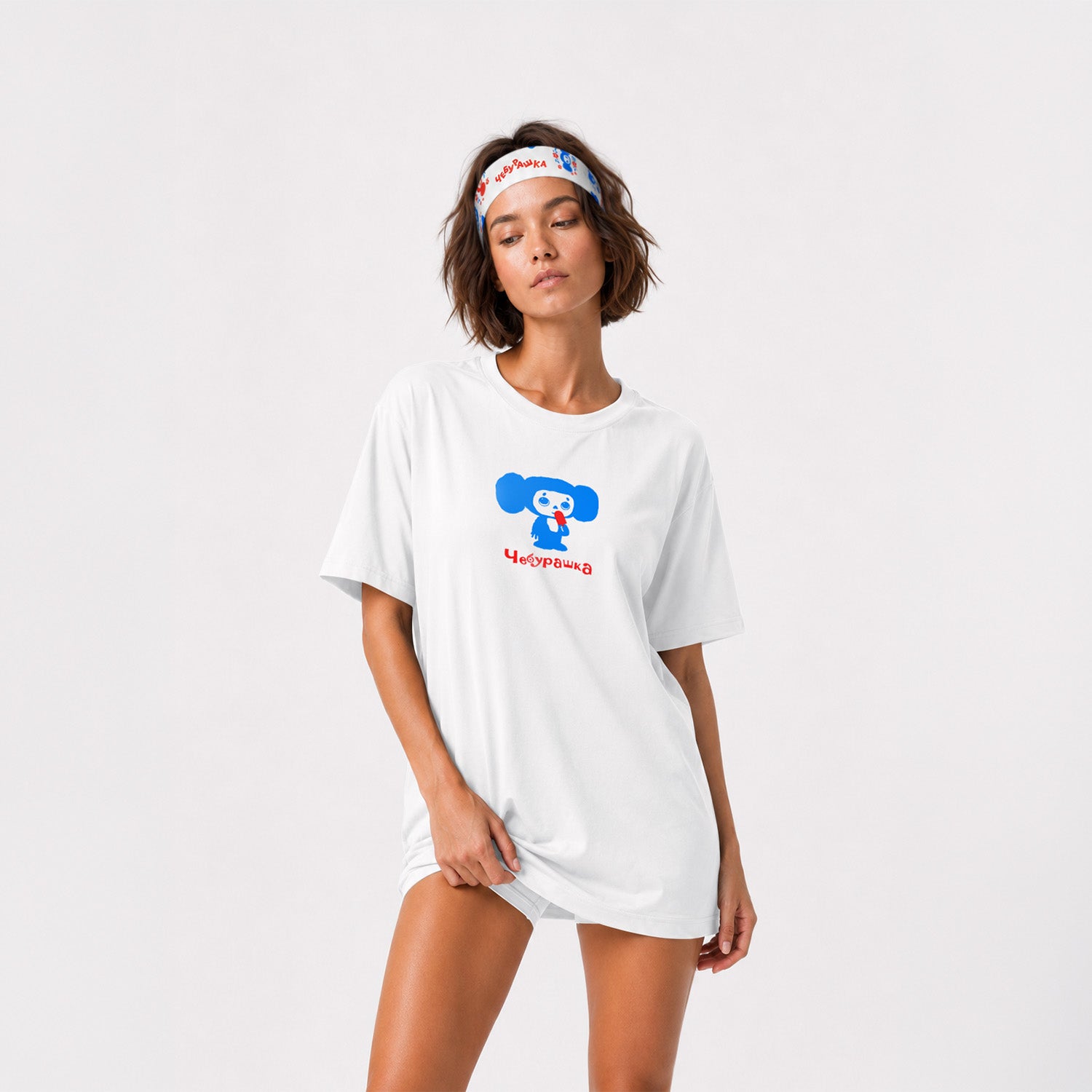 A cute fit model girl wearing white athletic headband featuring a colorful Soviet cartoon character Cheburashka pattern in blue and red, with the brand name 'Чебурашка' printed in black on the inside. Front side.