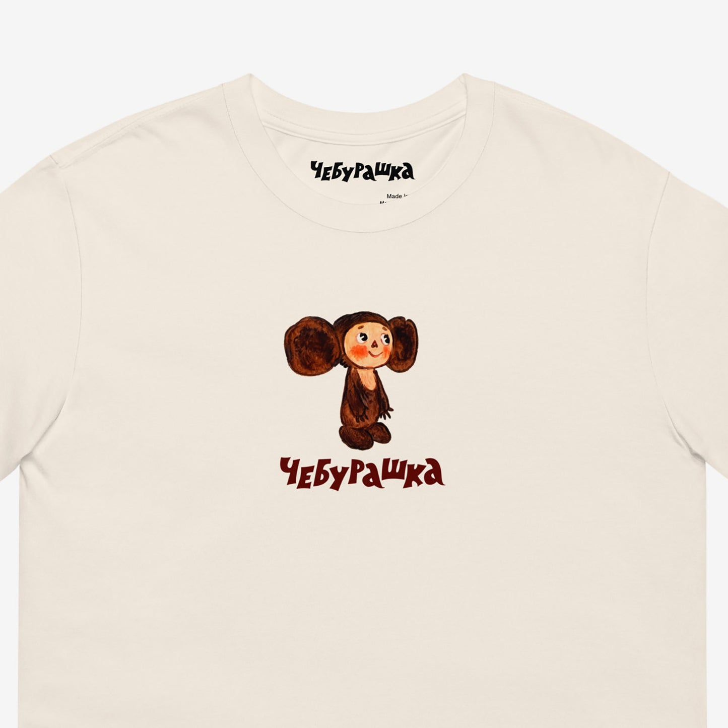Unisex natural-white Tee featuring original Soviet cartoon character clumsy painted Cheburashka and Чебурашка logo in brown. High-quality 100% cotton. Closeup.