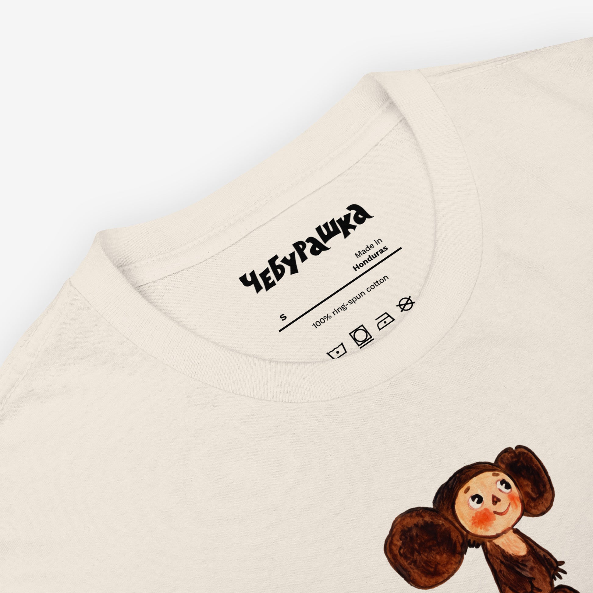Unisex natural-white Tee featuring original Soviet cartoon character clumsy painted Cheburashka and Чебурашка logo in brown. High-quality 100% cotton. Closeup necktag.