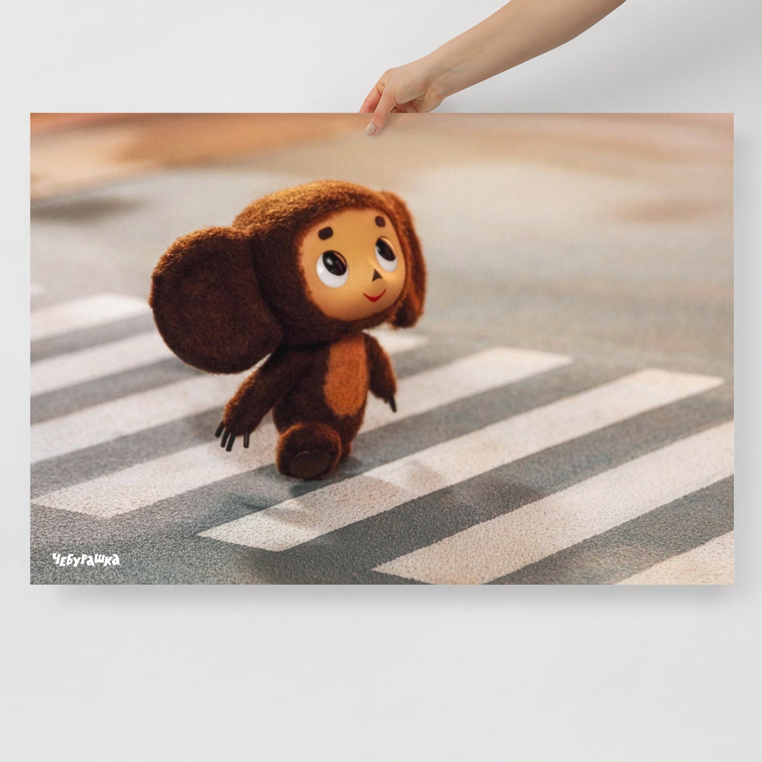 Hand holding large 91x61cm poster with Cheburashka on road zebra crossing. Still frame from old original Soviet cartoon. With logo Чебурашка.