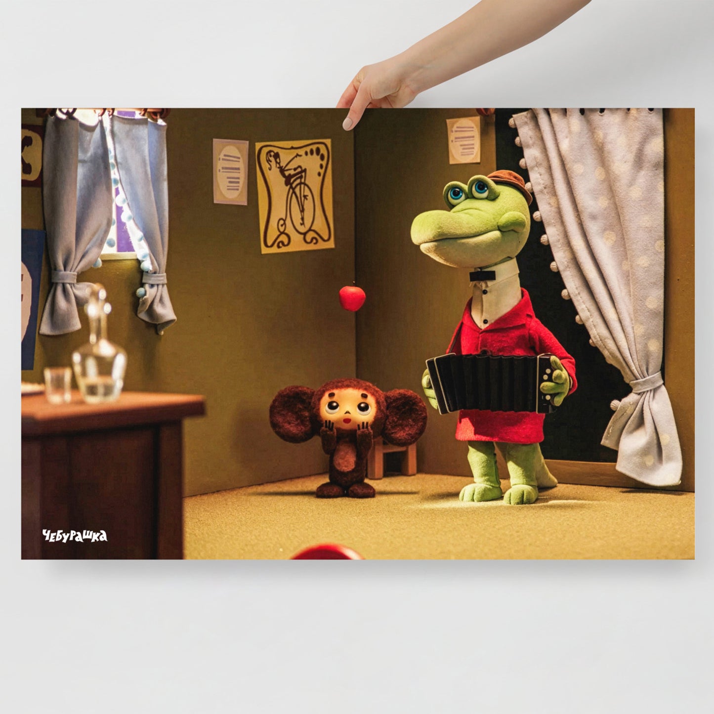 A hand holding large art print featuring a scene from the Soviet cartoon Cheburashka, showing Cheburashka playing with apples and his friend Gena the crocodile playing accordion in a room. With Чебурашка logo on the bottom left side.
