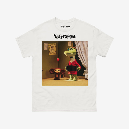 White unisex T-shirt featuring a scene from the Soviet cartoon Cheburashka, showing Cheburashka playing with apples and his friend Gena the crocodile playing accordion in a room. With Чебурашка logo above the image.
