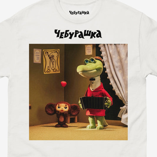White unisex T-shirt featuring a scene from the Soviet cartoon Cheburashka, showing Cheburashka playing with apples and his friend Gena the crocodile playing accordion in a room. With Чебурашка logo above the image. Closup.