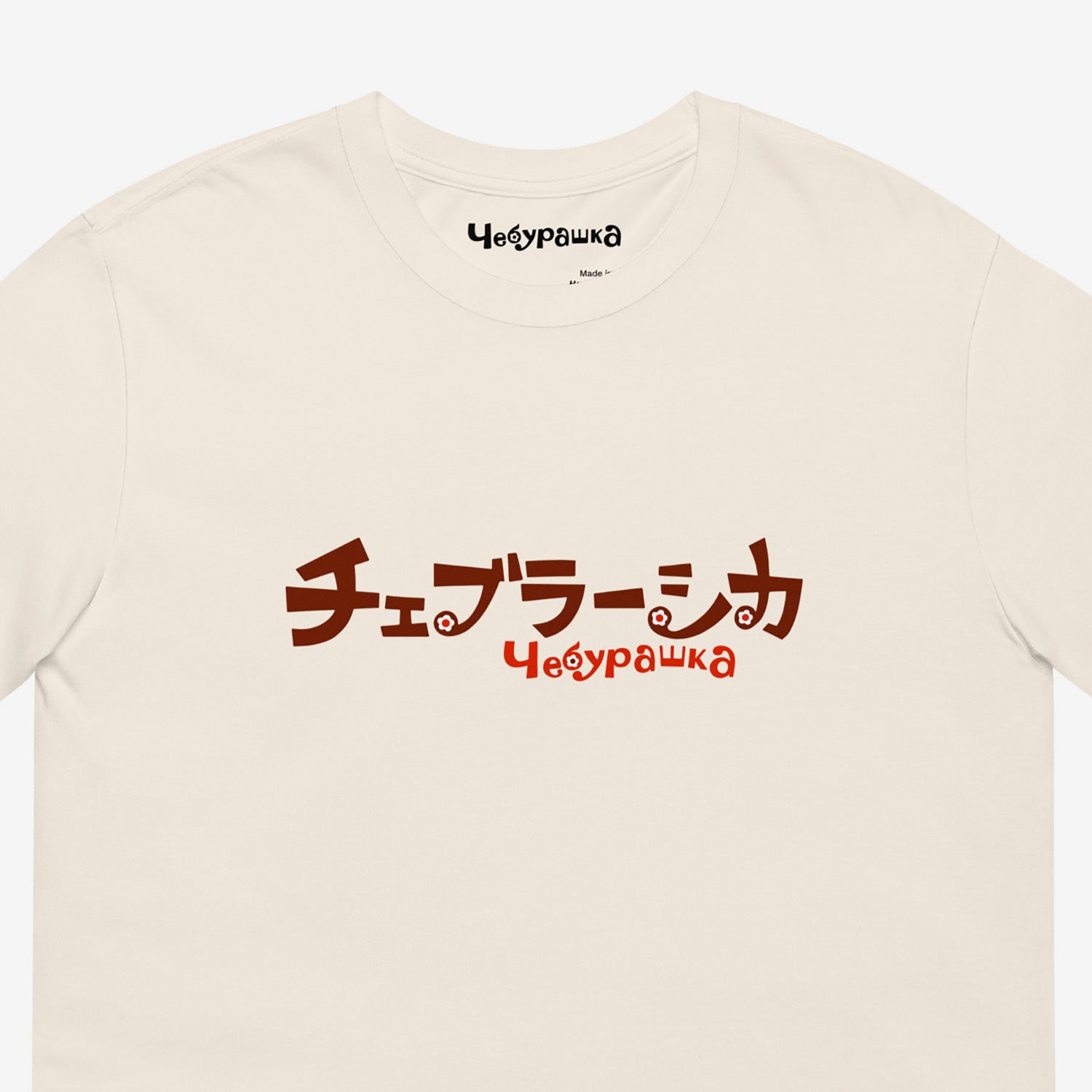 A beige short-sleeve unisex Cheburashka, original Soviet cartoon Tee with a brown チェブラーシカ logo in japanese and a smaller 'Чебурашка' logo in russian underneath. Closeup.