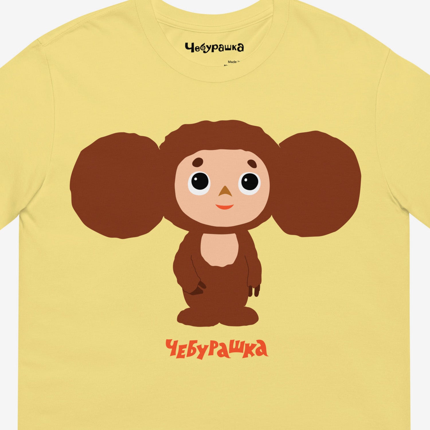 Yellow unisex adult T-shirt featuring Soviet cartoon shy Cheburashka. Minimalist large bright print design. With Чебурашка logo below and on the necktag. Closeup photo.