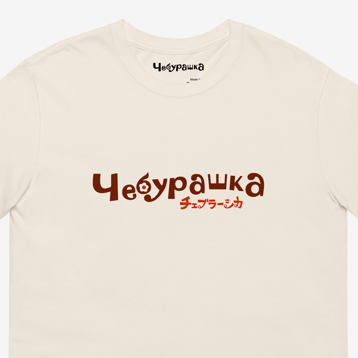 A beige short-sleeve unisex Cheburashka, original Soviet cartoon Tee with a brown 'Чебурашка' logo in russian and smaller underneath logo in japanese チェブラーシカ. Closeup print details.