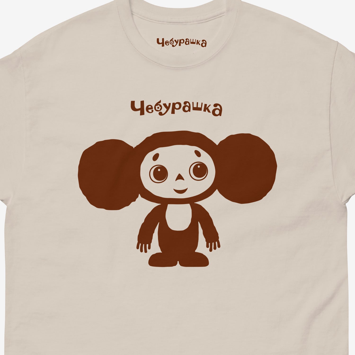 Beige unisex adult T-shirt featuring Soviet cartoon Cheburashka, standing and smiling. Minimalist one-color design. With Чебурашка logo above the image. Closeup photo.