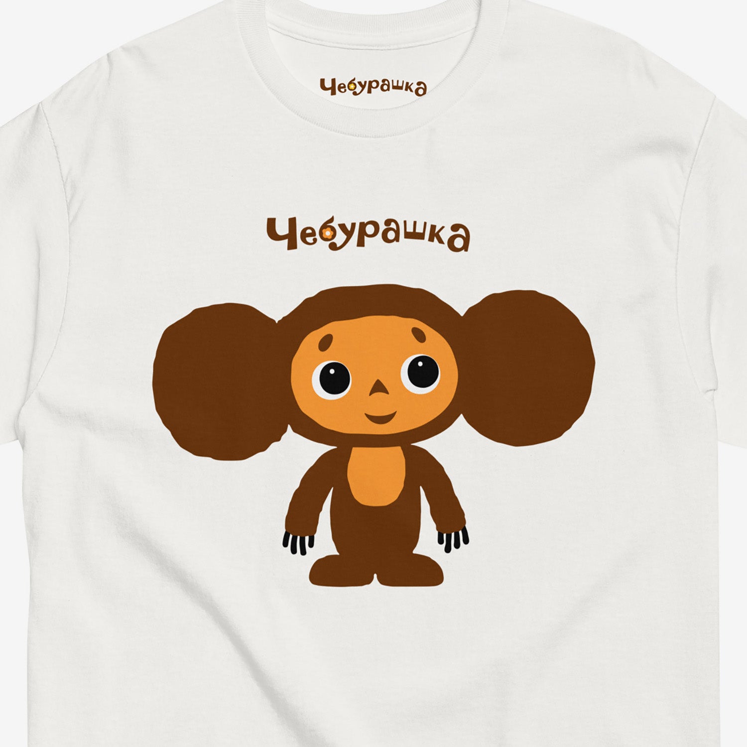 White unisex T-shirt featuring Soviet cartoon Cheburashka, standing and smiling. With Чебурашка logo above the image. Closeup photo.