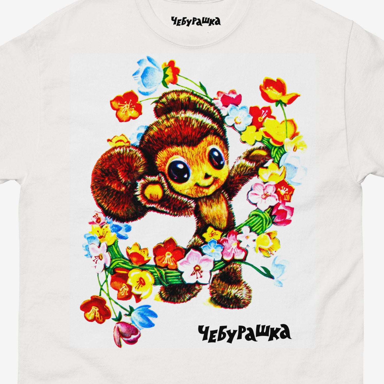 White unisex T-shirt with large print of frolicking Cheburashka painted with flowers, from Soviet cartoon. With logo Чебурашка. Closeup photo.