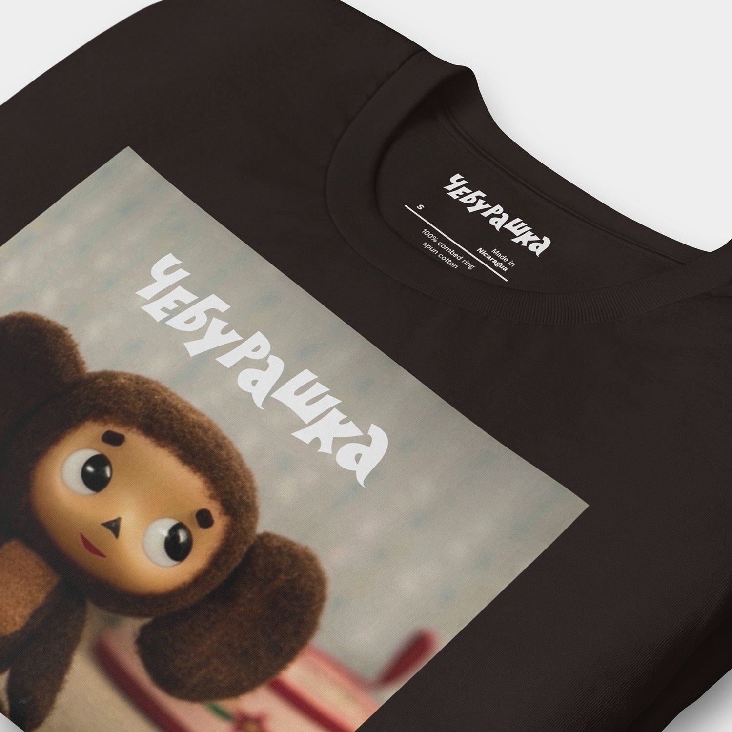Brown unisex T-shirt with large print of original Soviet cartoon character Cheburashka standing cute and curious. Featuring Чебурашка logo at the of the print. Closeup print details.