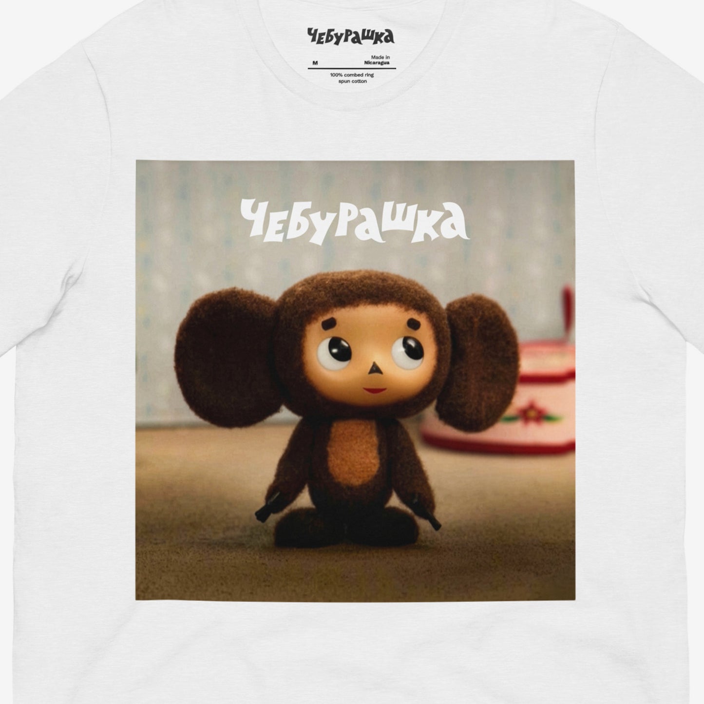 White unisex T-shirt with large print of original Soviet cartoon character Cheburashka standing cute and curious. Featuring Чебурашка logo at the of the print. Front closeup.