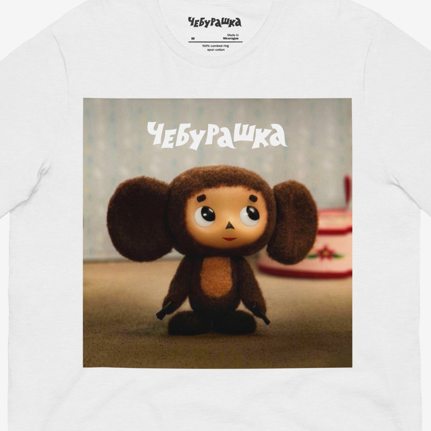 White unisex T-shirt with large print of original Soviet cartoon character Cheburashka standing cute and curious. Featuring Чебурашка logo at the of the print. Front closeup.