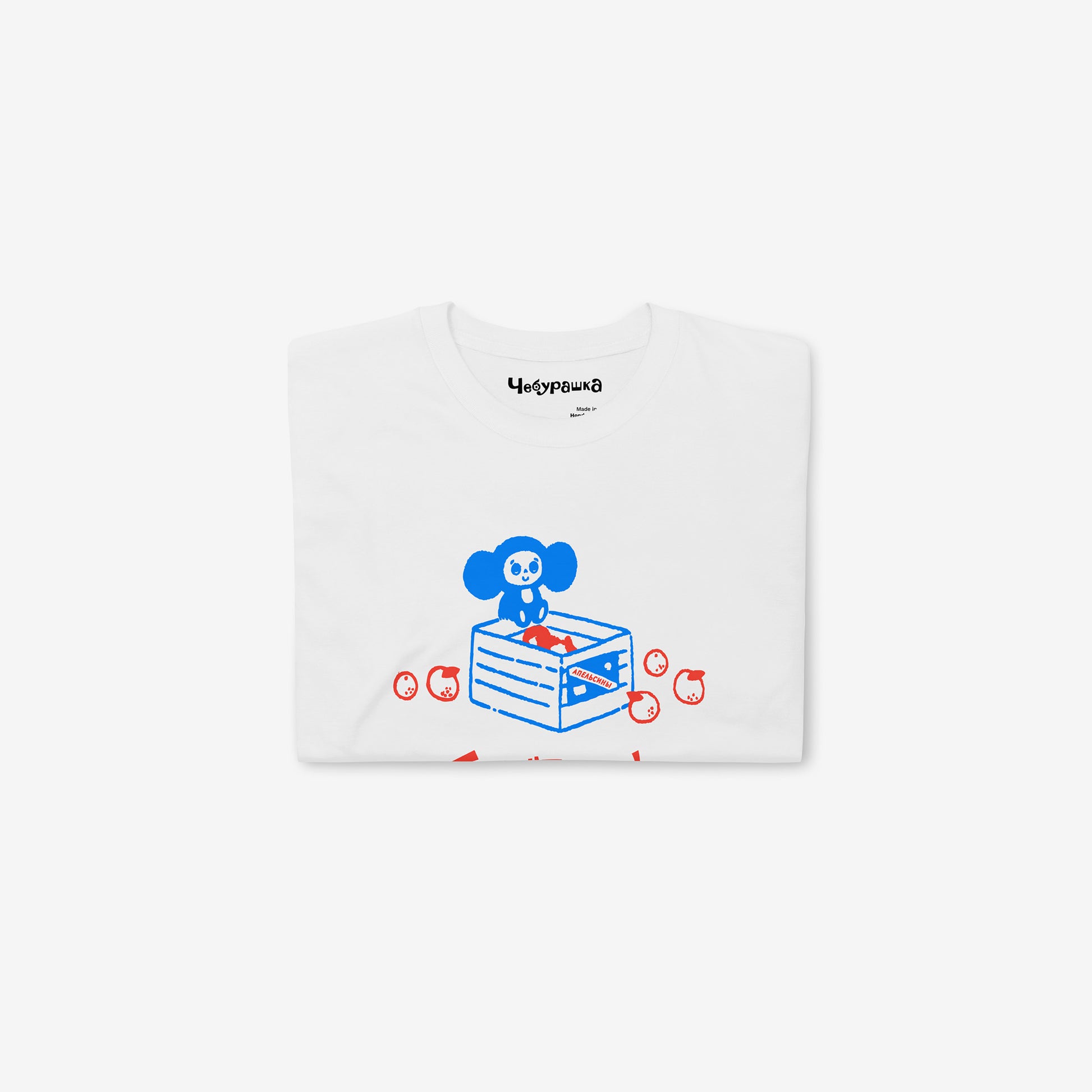 White unisex T-shirt with original Soviet cartoon character Cheburashka sitting on a box of oranges and チェブラーシカ logo in red. Folded.