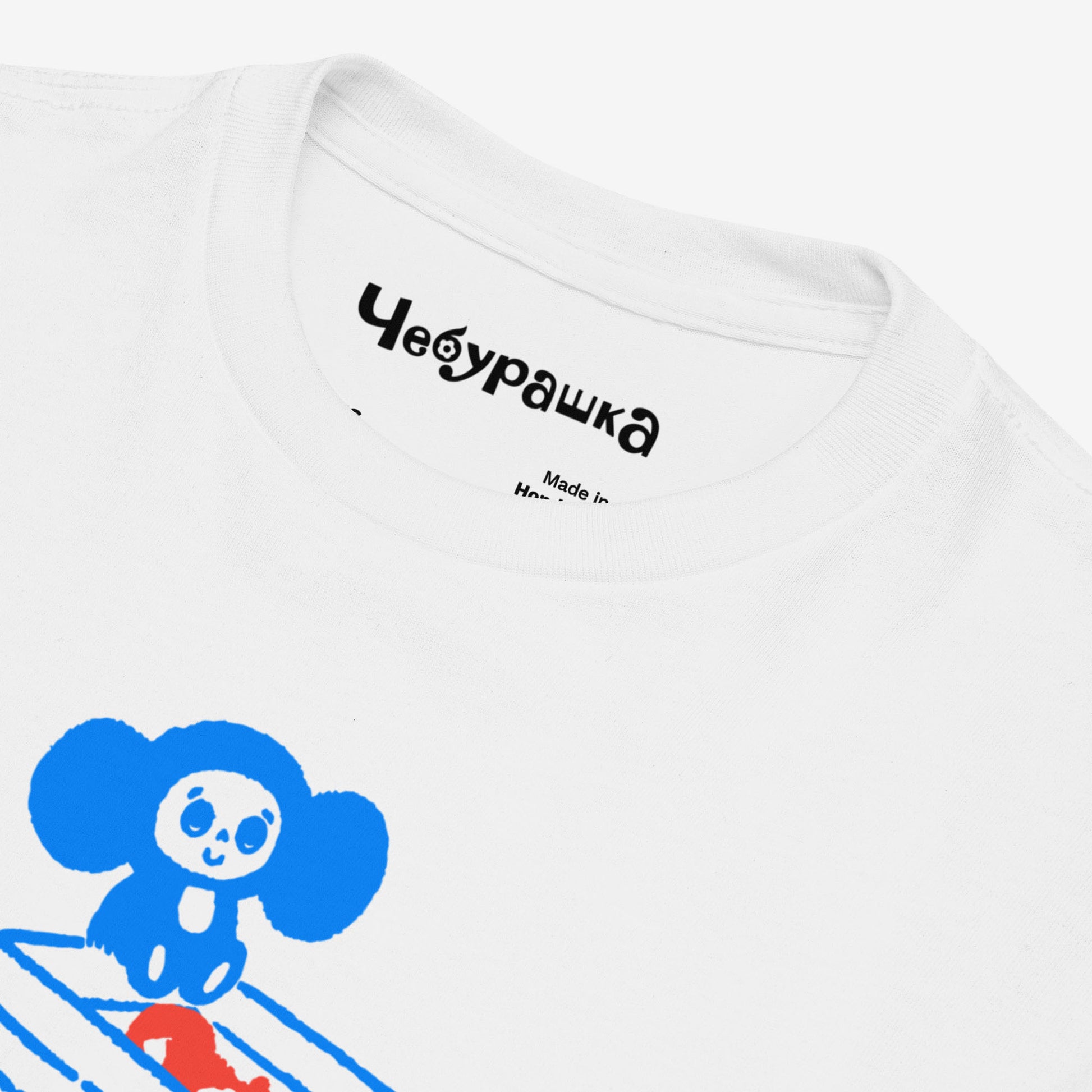 White unisex youth T-shirt with original Soviet cartoon character Cheburashka sitting on a box of oranges and チェブラーシカ logo in red. Front closeup.
