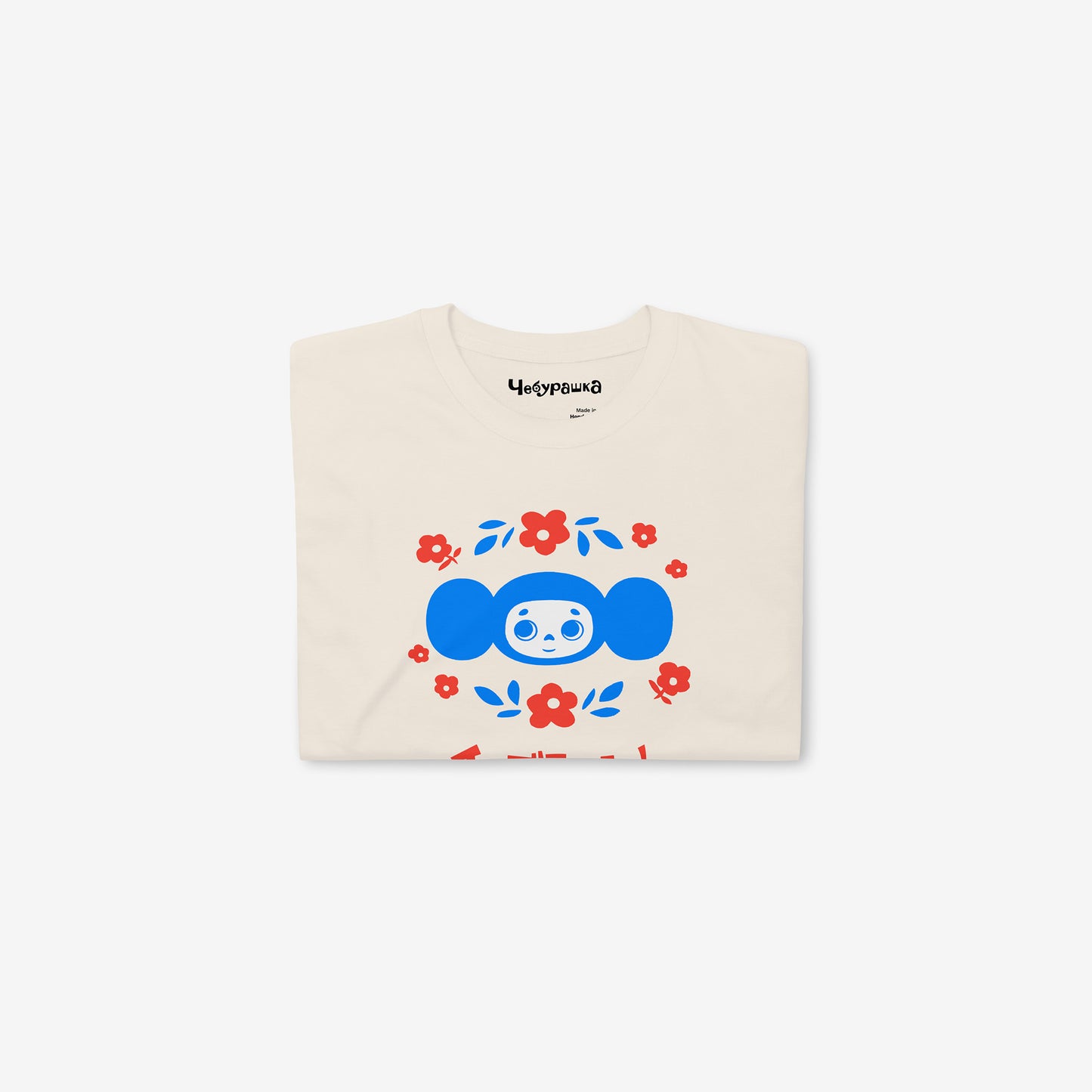 Beige unisex T-shirt with original Soviet cartoon character Cheburashka among a pattern of flowers and with logo in japanese チェブラーシカ. Folded.