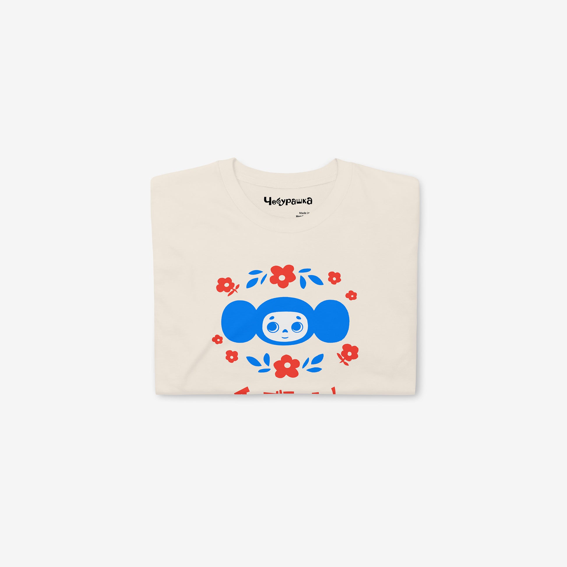 Beige unisex T-shirt with original Soviet cartoon character Cheburashka among a pattern of flowers and with logo in japanese チェブラーシカ. Folded.