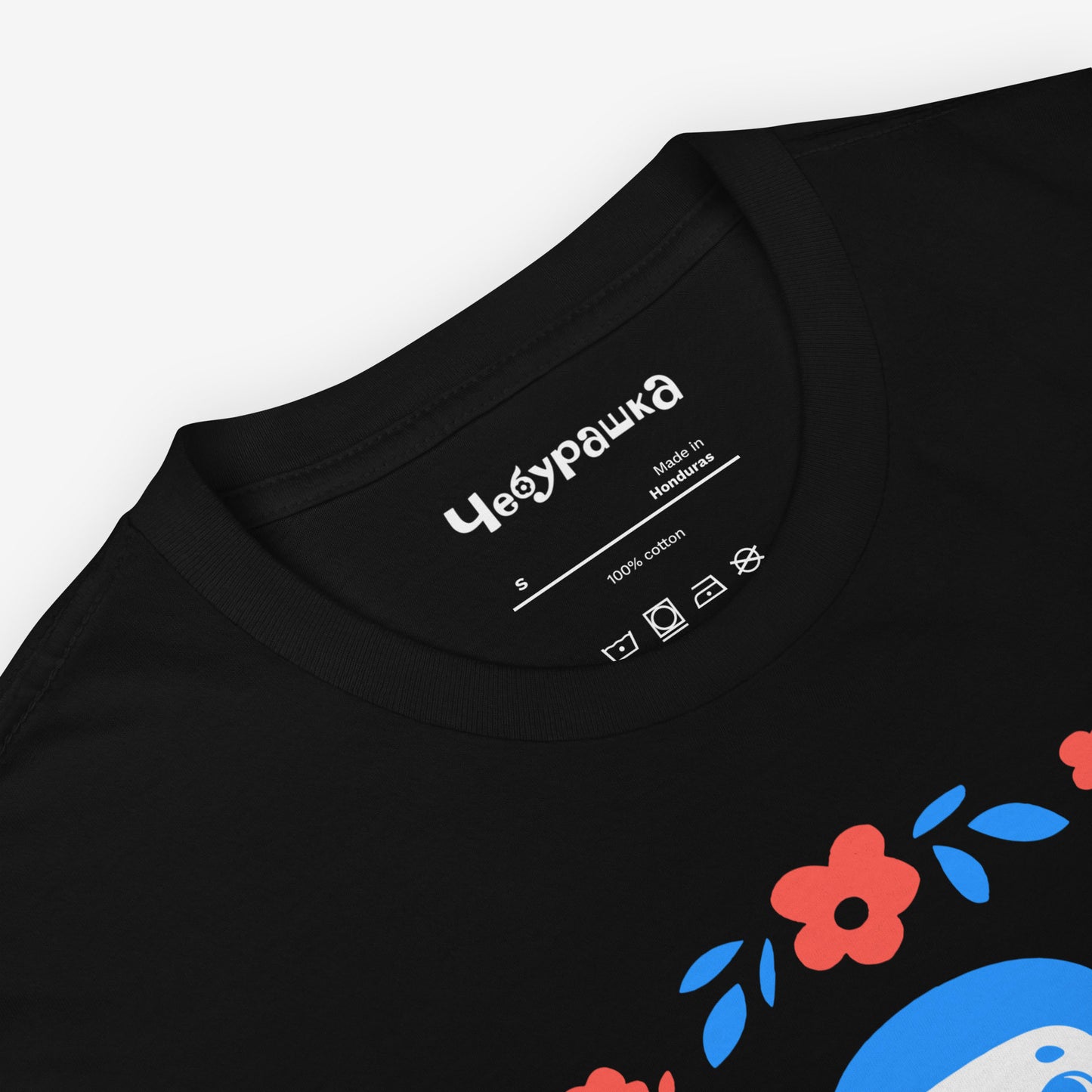 Black unisex T-shirt with original Soviet cartoon character Cheburashka among a pattern of flowers and with logo in japanese チェブラーシカ. Closeup print details.
