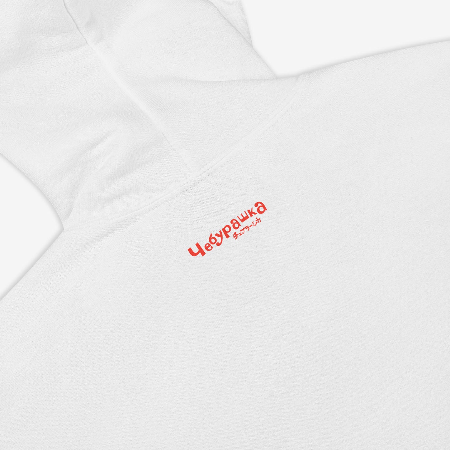 White unisex hoodie with minimalist graphic of original smiley Soviet cartoon character Cheburashka and チェブラーシカ logo in red. Back side with small Чебурашка logo. Closeup photo.