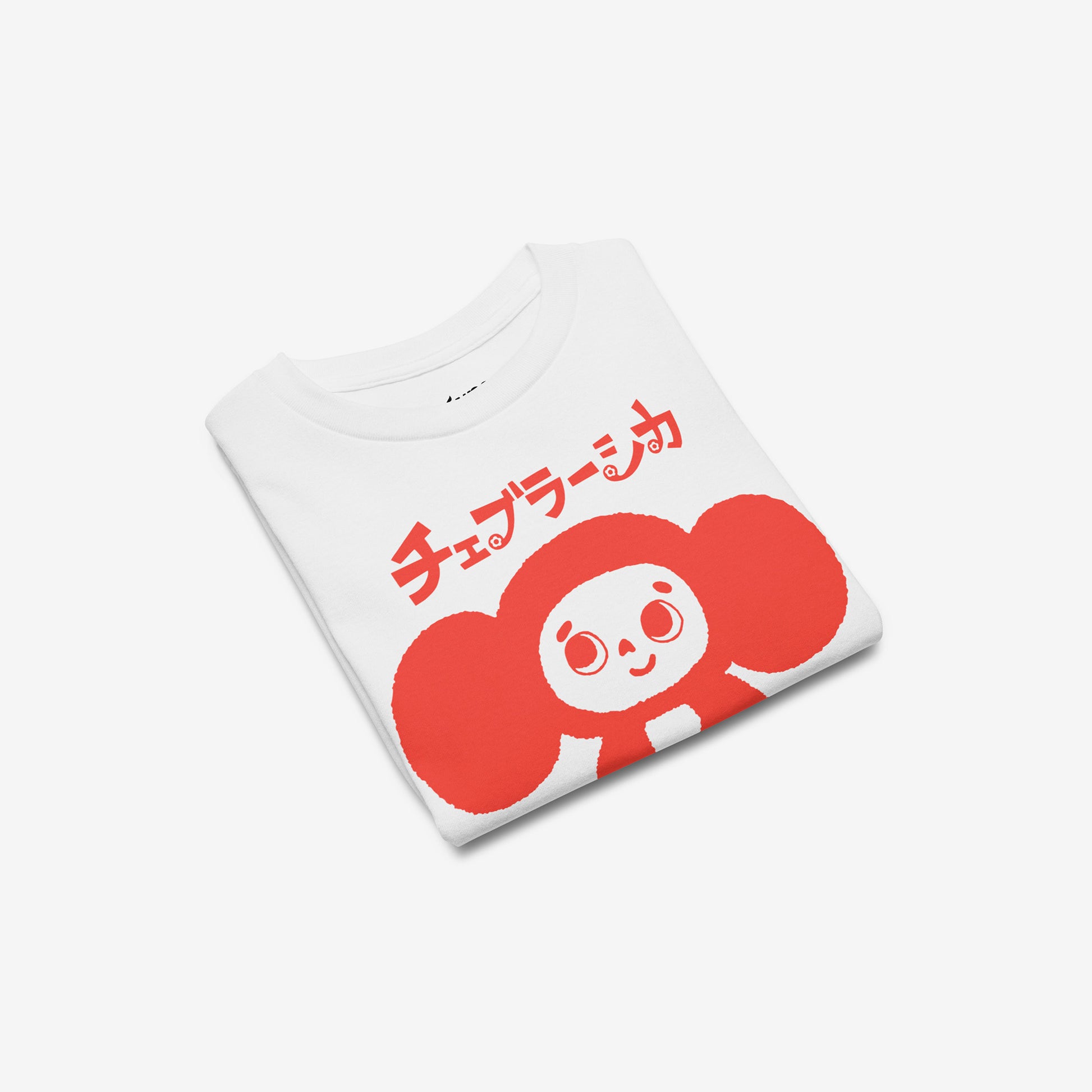 White unisex youth T-shirt with minimalist graphic of original smiley Soviet cartoon character Cheburashka and チェブラーシカ logo in red. Folded.
