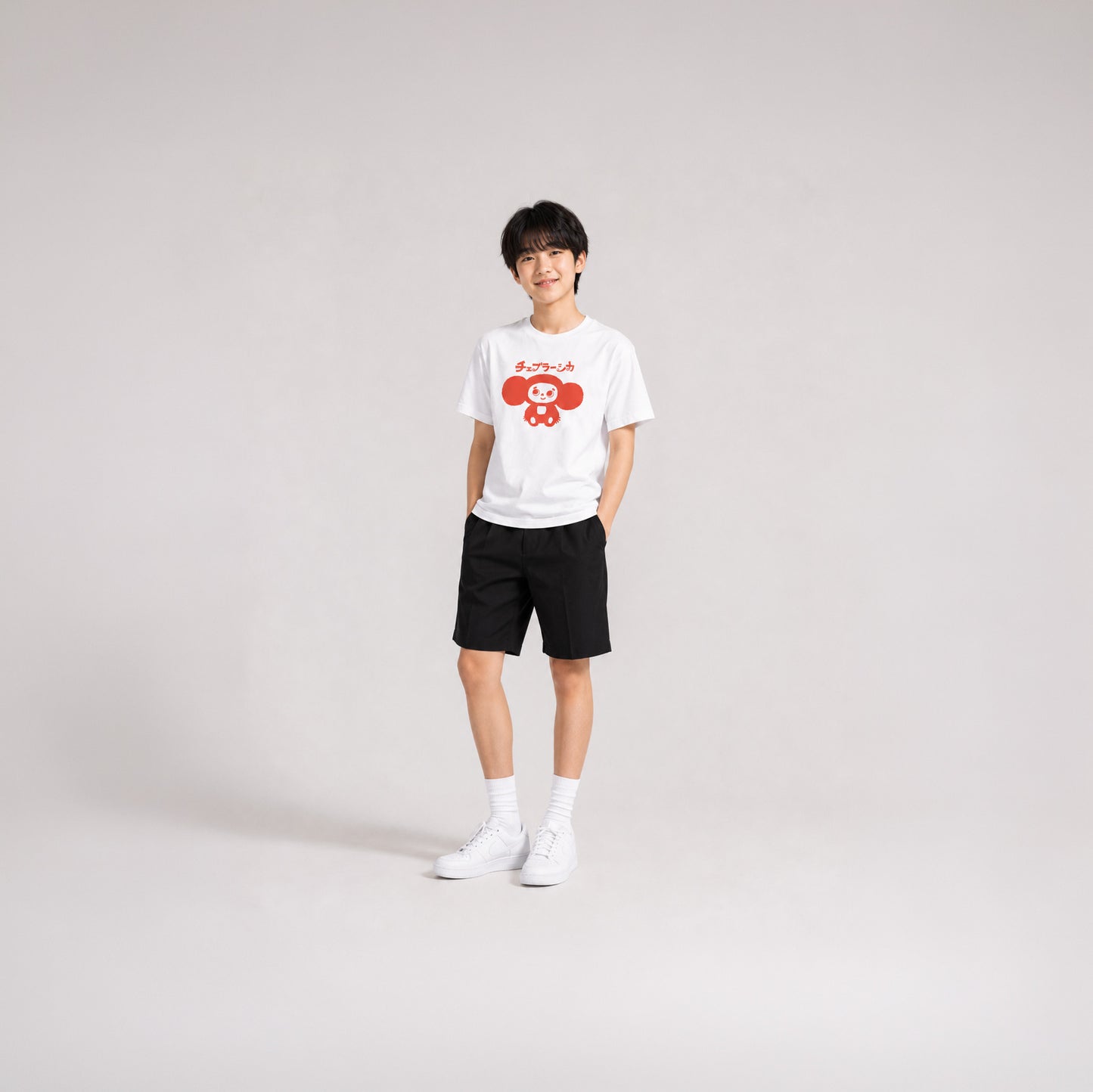Japanese school boy model wearing shorts and white unisex youth T-shirt with minimalist graphic of original smiley Soviet cartoon character Cheburashka and チェブラーシカ logo in red.