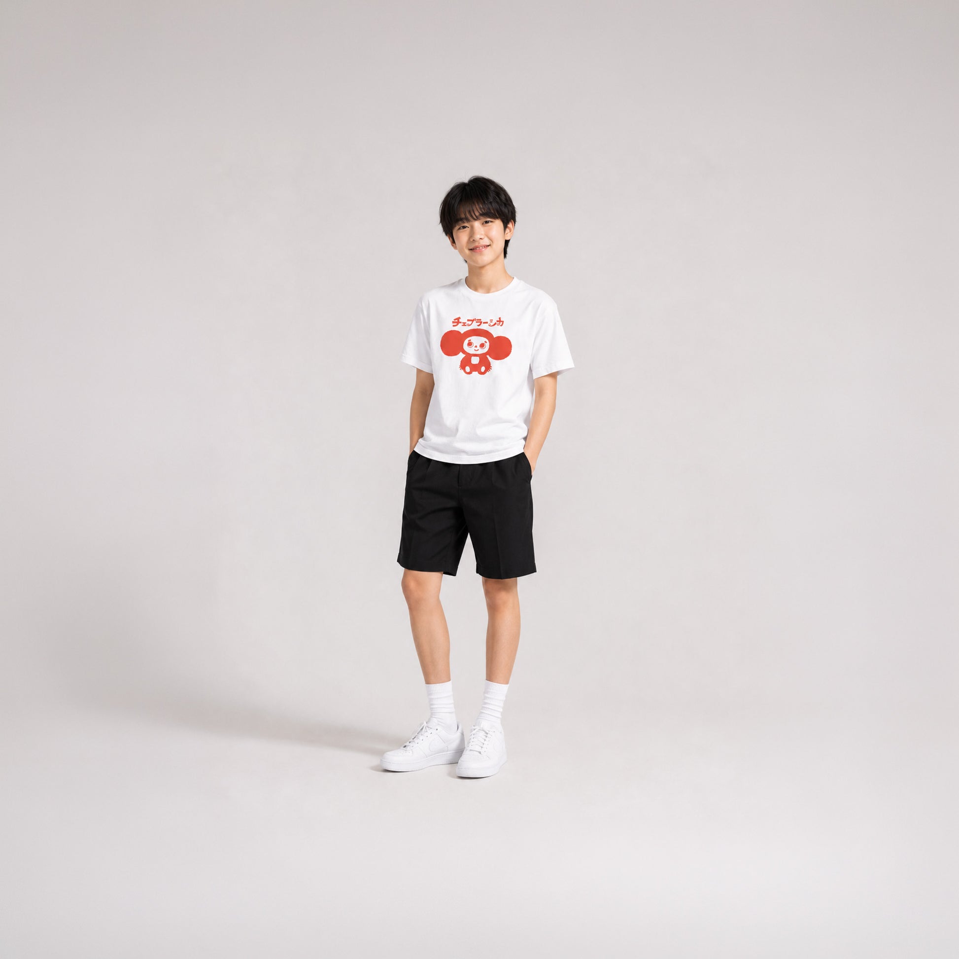 Japanese school boy model wearing shorts and white unisex youth T-shirt with minimalist graphic of original smiley Soviet cartoon character Cheburashka and チェブラーシカ logo in red.