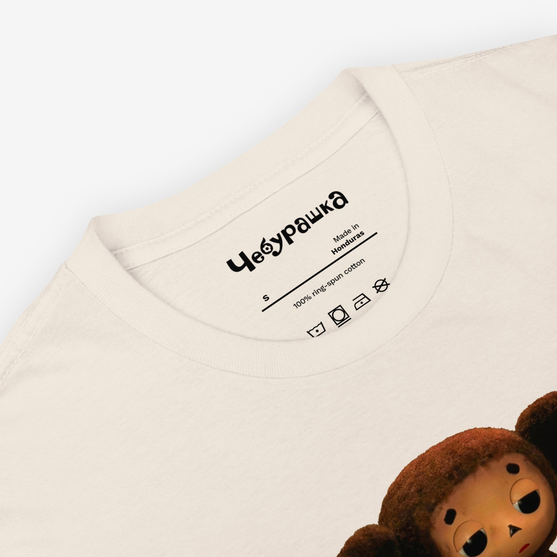 Beige unisex T-shirt featuring large print of Sad Cheburashka from Soviet cartoon. With logo Чебурашка. Closeup.