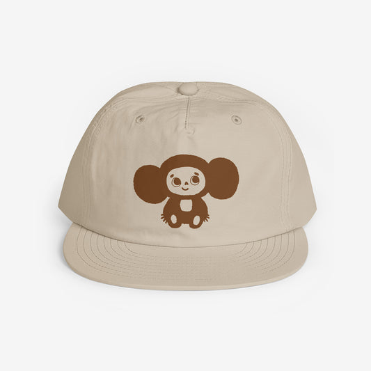 A beige surf cap with a flat brim, featuring brown Soviet cartoon character Cheburashka on the front. Front side.