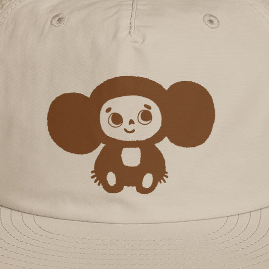 A beige surf cap with a flat brim, featuring brown Soviet cartoon character Cheburashka on the front. Closeup.